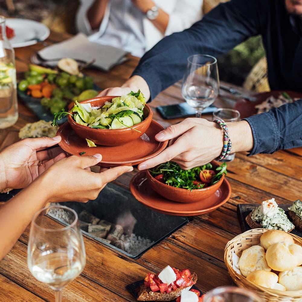 A group of people seated at a restaurant, two people pass a bowl of salad across the table