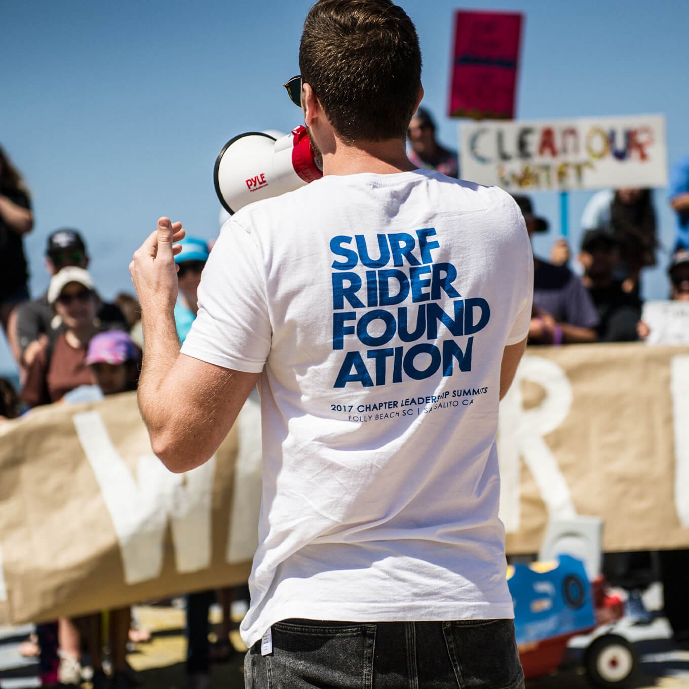 Man wearing Surfrider shirt with megaphone faces crowd of activists.